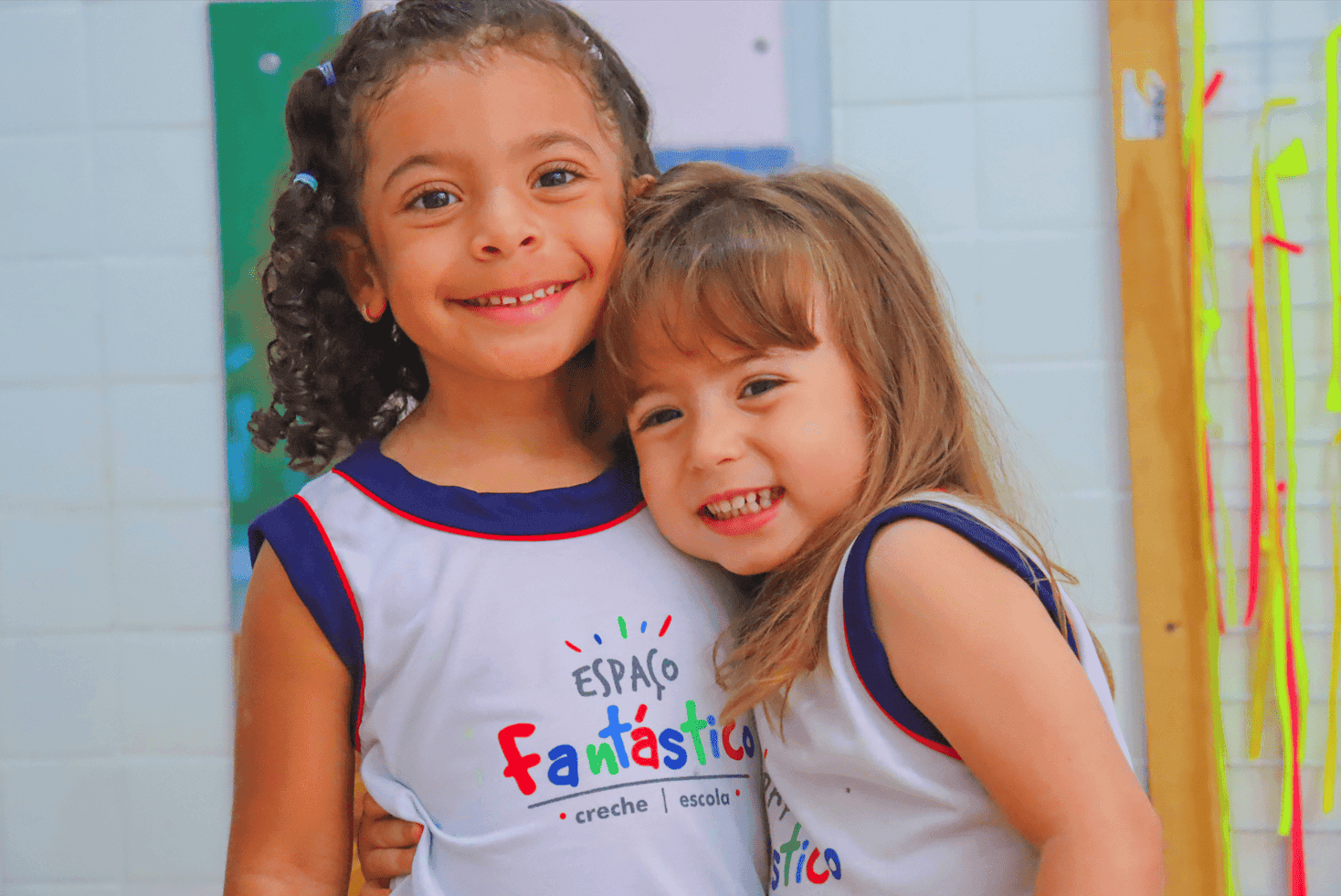 Duas meninas sorridentes, usando uniformes da escola 'Espaço Fantástico', se abraçam em um ambiente escolar colorido e alegre.