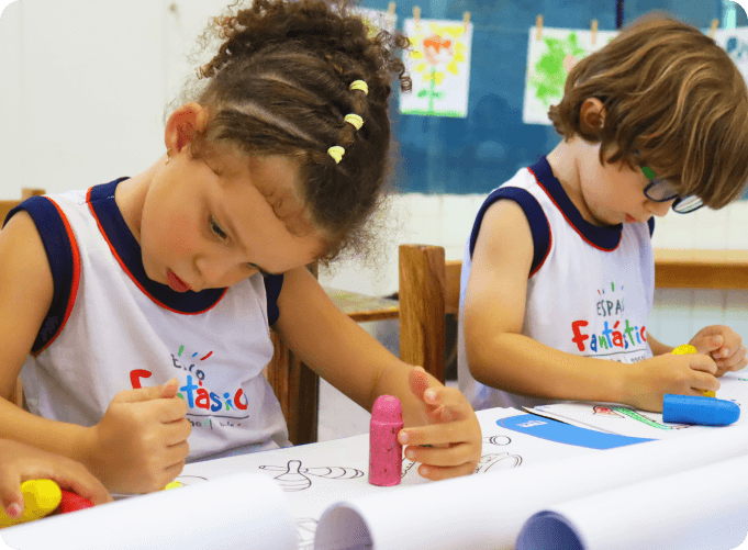 Crianças pequenas desenhando e colorindo em sala de aula, concentradas em suas atividades artísticas, usando uniformes do Espaço Fantástico.