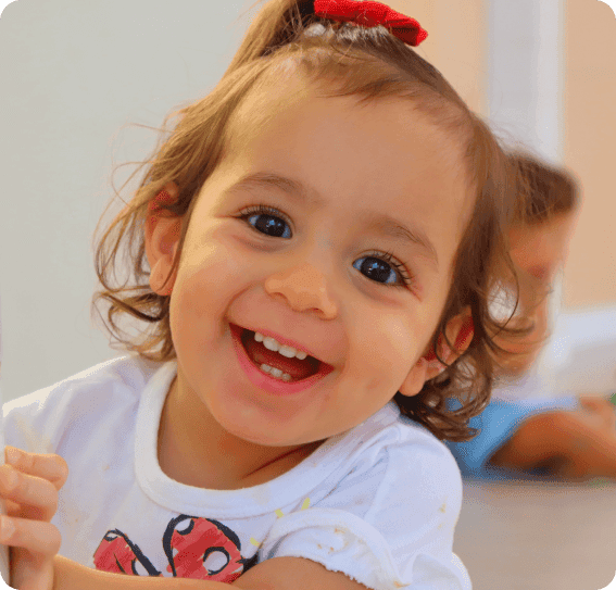 Criança sorridente com laço vermelho no cabelo, usando camiseta branca, em ambiente iluminado.