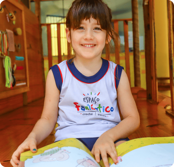 Criança sorridente, sentada no chão de uma sala de aula colorida, vestindo uniforme branco e azul com o logotipo “Espaço Fantástico – creche e escola”, enquanto segura um livro infantil aberto.