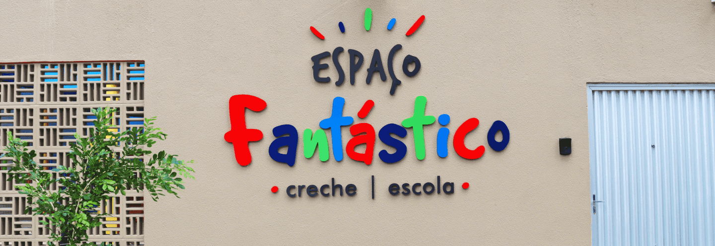 Fachada do Espaço Fantástico, creche e escola, com o nome colorido em letras grandes e divertidas sobre uma parede bege. Há uma planta verde à esquerda e uma porta metálica à direita.